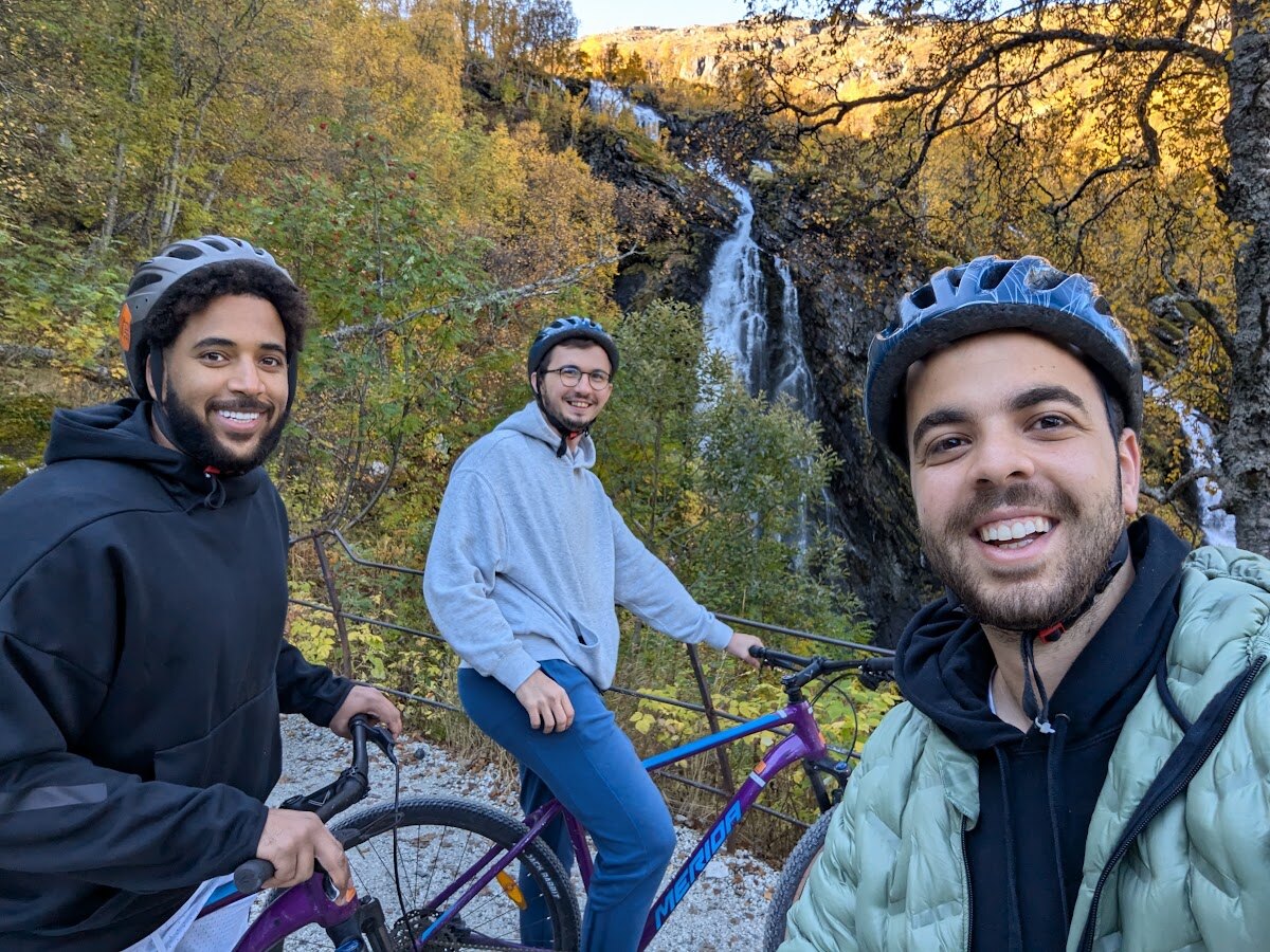 Three cyclists smiling with their mountain bikes in front of a waterfall in Flåm valley